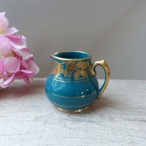 Op de afbeelding: Een klein, tealblauw keramiek melkkannetje met gouden rand en bloemmotieven. Het melkkannetje heeft een rond lichaam en een klein handvat.
