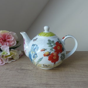 Peut inclure: Théière en porcelaine blanche avec un motif floral composé de fleurs rouges, orange, jaunes, bleues et vertes et de papillons. La théière a un couvercle vert et une anse.