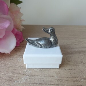 Peut inclure: Une figurine de canard en argent posée sur une boîte blanche. Le canard est tourné vers la droite de l'image et présente un design détaillé.