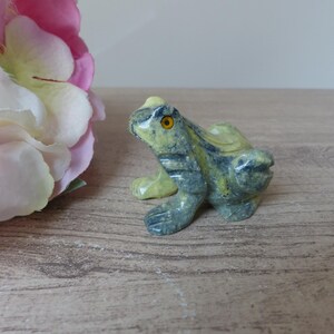 Könnte beinhalten: Eine kleine Froschfigur aus geschnitztem Stein mit gesprenkelten Grün- und Gelbtönen und einer glatten, polierten Oberfläche. Der Frosch hat ein markantes, rundes orangefarbenes Auge und ist in einer sitzenden, wachsamen Haltung dargestellt.