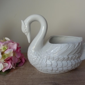 Può includere: Un vaso in ceramica bianca a forma di cigno con un lungo collo curvo e dettagliate texture di piume modellate sul corpo. Il cigno presenta una cavità superiore aperta per contenere oggetti ed è posizionato accanto a un mazzo di ortensie rosa e bianche.