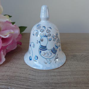 Può includere: Campana in ceramica bianca con un disegno dipinto a mano raffigurante un uccello nei toni del blu e dell'oro. La campana ha una base svasata e una parte superiore arrotondata, con dettagli floreali e a spirale. La campana è alta circa 13 cm.