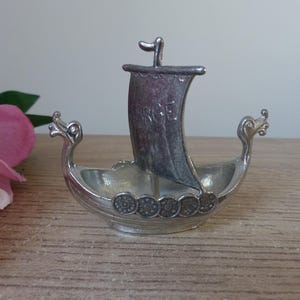 Könnte beinhalten: Eine silberne Zinn-Figur eines Wikinger-Schiffes mit einem Segel, auf dem "SROE" steht. Das Schiff hat an jedem Ende einen Drachenkopf und eine Reihe runder Verzierungen am Boden.