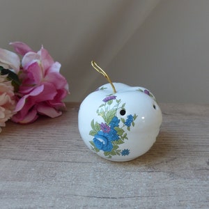 Może przedstawiać: Biały ceramiczny jabłko z kwiatowym wzorem w odcieniach niebieskiego, zielonego i fioletowego. Jabłko ma przymocowaną złotą sznurówkę.