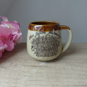 Könnte beinhalten: Eine braune und beige Keramiktasse mit braunem Rand und Henkel. Die Tasse hat ein Weizenmuster und den Text "Live Better Naturally" auf der Vorderseite.