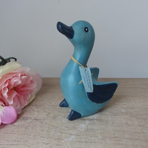 Puede incluir: Una figurita de pato de cerámica azul con pico y ojos negros. El pato está de pie sobre una superficie de madera. La figurita tiene una etiqueta adjunta que dice "Petites trouvailles de la mer" y "Calvados".