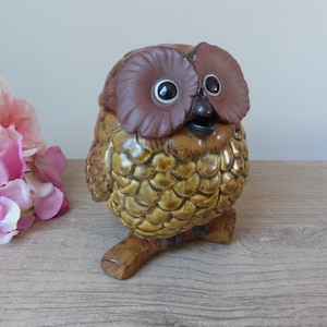 Peut inclure: Une figurine de hibou en céramique brune et jaune avec de grands yeux et un corps texturé. Le hibou est perché sur deux petits pieds.