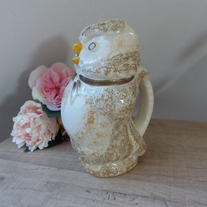 Può includere: Brocca in ceramica a forma di gufo, con base bianca e dettagli maculati marroni. Il gufo ha becco giallo, colletto marrone e occhi aperti. Fiori rosa visibili sullo sfondo. Un oggetto decorativo.