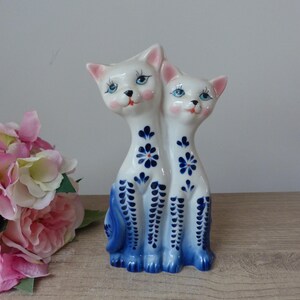 Könnte beinhalten: Eine weiße Keramik-Katzenfigur mit zwei verbundenen Körpern. Die Katzen haben blaue Augen, rosa Ohren und rosige Wangen. Blaue Blumen- und Musterdesigns zieren die Körper und Schwänze. Der Sockel und die Details sind in leuchtendem Blau bemalt.