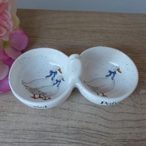 Può includere: Saliera e pepe in ceramica bianca con una glassa macchiettata. Ogni saliera presenta un disegno dipinto a mano di tre oche bianche con nastri blu. Le saliere sono collegate da un manico e hanno le parole "Sel" e "Poivre" scritte sul fondo.