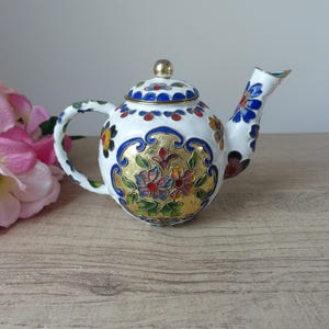 Könnte beinhalten: Eine weiße Teekanne mit goldenem Blumenmuster. Die Teekanne hat einen runden Körper und einen kleinen Ausguss. Das Blumenmuster ist in Blautönen, Rot, Grün und Gold gehalten.