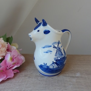 Puede incluir: Una jarra de cerámica azul y blanca con forma de vaca con un molino de viento y casas pintadas en el lateral.