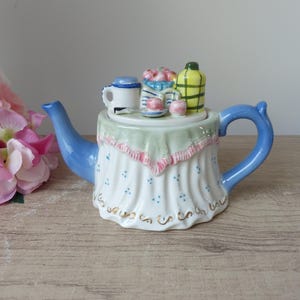 Può includere: Una teiera decorativa in ceramica con manico e beccuccio blu. La teiera è bianca con un design blu e oro. Il coperchio presenta oggetti in miniatura in ceramica, tra cui una tazza, una bottiglia e un piattino con oggetti rosa.