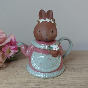 Può includere: Una teiera in ceramica con coperchio a forma di coniglietto. La teiera è rosa e blu, con un coniglietto che indossa una corona floreale e tiene un cestino. La teiera ha un manico e un beccuccio. Il coniglietto è marrone.