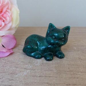 Könnte beinhalten: Eine kleine, grüne Keramik-Katzenfigur. Die Katze liegt mit dem Kopf zur Seite gedreht. Die Figur hat eine glänzende Oberfläche.