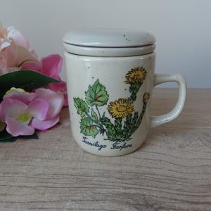 May include: Cream-colored ceramic mug with a lid, featuring a botanical illustration of yellow flowers and green leaves. The mug has a handle and the text "Tussilago Farfara" is printed on the front.