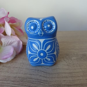 Może przedstawiać: Niebieska ceramiczna figurka sowy z białymi wzorami kwiatowymi i geometrycznymi. Sowa ma duże, okrągłe oczy i stylizowany design. Figurka stoi na drewnianej powierzchni, z różowymi i białymi kwiatami w tle.
