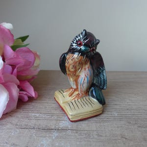 Op de afbeelding: Een keramiek uilbeeldje met zwarte, bruine, blauwe en witte verf, gepercht op een gesloten boek met een rode rug. De uil heeft rode ogen en kijkt naar links.