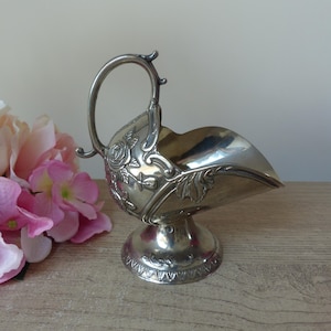 Op de afbeelding: Zilveren suikerpot met een handvat en bloemendetails. De kom heeft een gebogen ontwerp en staat op een decoratieve voet. De handgreep is gebogen en de kom is leeg. De achtergrond bevat roze en perzik bloemen.