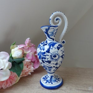 Op de afbeelding: Een wit en blauw keramieken vaas met een decoratieve handgreep. De vaas heeft een bloemmotief in blauw tegen een witte achtergrond. De voet en de rand zijn ook blauw. De vaas staat op een houten oppervlak met roze bloemen op de achtergrond.