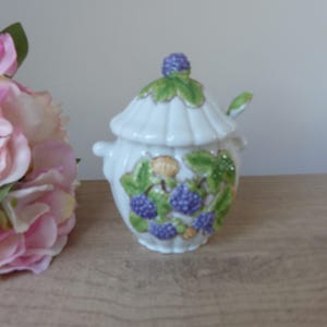 Puede incluir: Un azucarero de cerámica blanca con tapa y una cucharita. El cuenco está decorado con moras moradas, hojas verdes y bayas amarillas. La tapa está rematada con una mora morada y hojas verdes.