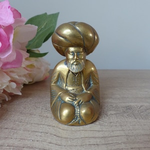 Può includere: Una statuetta in ottone di un uomo seduto con un turbante e una lunga barba. La figura ha le mani giunte in grembo. La statuetta è su una superficie di legno con fiori rosa e bianchi sullo sfondo.