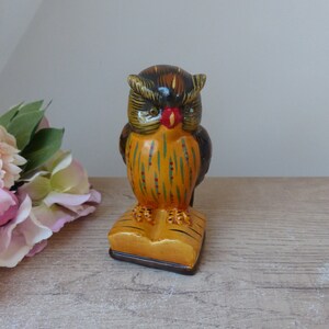 May include: A ceramic owl figurine with black, brown, yellow, green, and red paint. The owl is perched on a stack of books with a brown base.