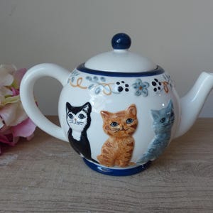 Può includere: Una teiera in ceramica bianca con base, manico e pomello blu. La teiera presenta tre illustrazioni di gatti in nero, arancione e blu. Disegni floreali e di impronte di zampe decorano il bordo superiore. La teiera è su una superficie di legno.