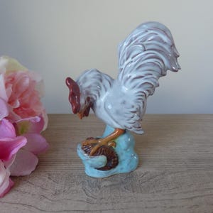 Può includere: Una statuetta di gallo in ceramica bianca con cresta e bargigli marroni, in piedi su una base blu e marrone. Il gallo è rivolto a sinistra e ha le penne della coda sollevate.