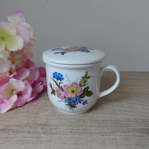 Peut inclure: Tasse à thé en céramique blanche avec un motif floral composé de fleurs roses, jaunes et bleues. La tasse à thé a un couvercle et une anse.