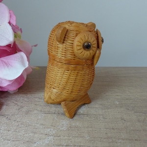 May include: A woven wicker owl figurine with brown eyes. The owl is standing on a wooden base.