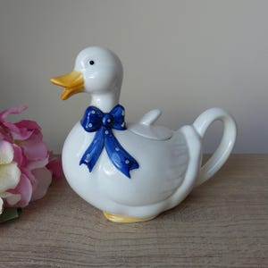Peut inclure: Une théière en céramique blanche en forme de canard. Le canard présente un bec et des pattes jaunes, un œil noir et un ruban bleu à pois blancs noué autour du cou. La théière comprend un couvercle amovible sur le dos et une anse incurvée.