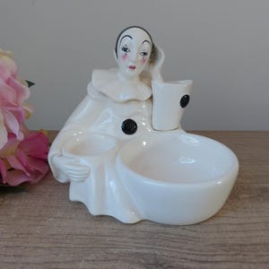 Peut inclure: Figurine de clown en céramique blanche avec un col à volants et des accents noirs. La figurine comporte un bol et une petite tasse attachés. Le visage du clown est peint avec des traits délicats, dont des joues rouges et une expression triste.