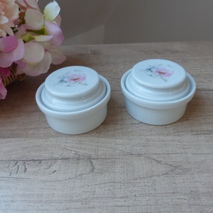 Puede incluir: Dos recipientes blancos de cerámica con tapa, adornados con un diseño floral en las tapas. Los recipientes son redondos y apilados, con una base ligeramente elevada. El diseño floral presenta acentos rosas y verdes. Sobre una superficie de madera marrón claro.