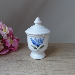 Może przedstawiać: Biały ceramiczny słoik z niebieskim wzorem kwiatu i pokrywką. Słoik ma podstawę na nóżkach.