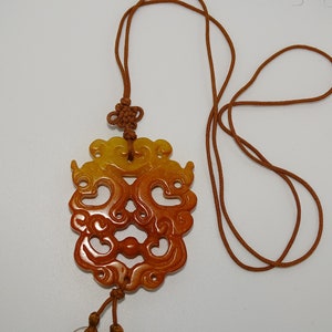 Op de afbeelding: Een bruine en gele jade hanger met ingewikkelde gravures, hangend aan een bruin koordsnoer.