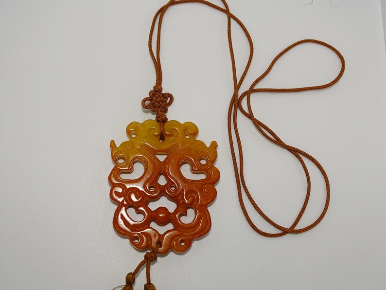 Op de afbeelding: Een bruine en gele jade hanger met ingewikkelde gravures, hangend aan een bruin koordsnoer.