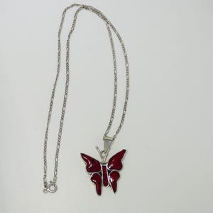 Op de afbeelding: Een zilverkleurige ketting met een vlinderhanger met een levendige rode emaille afwerking. De vlinderhanger is gedetailleerd met ingewikkelde vleugelontwerpen. De ketting is een delicate, geschakelde stijl, een klassiek sieraad.