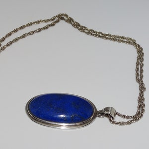 Op de afbeelding: Een zilveren ketting met een grote ovale hanger met een donkerblauwe lapis lazuli-steen.