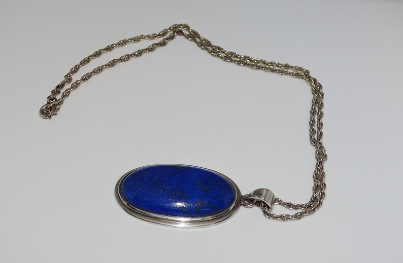 Op de afbeelding: Een zilveren ketting met een grote ovale hanger met een donkerblauwe lapis lazuli-steen.