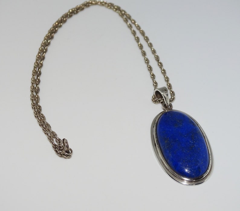 Op de afbeelding: Een zilveren ketting met een grote ovale hanger met een donkerblauwe lapis lazuli-steen.