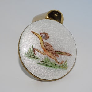 May include: A white enamel pendant with a gold-tone setting features a hand-painted image of a brown and yellow bird with green foliage. The bird is depicted in a running pose.
