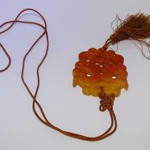 Op de afbeelding: Een gele en oranje jade hanger met ingewikkelde gravures hangt aan een bruin koord met een kwast. De hanger is met een knoop aan het koord gebonden.