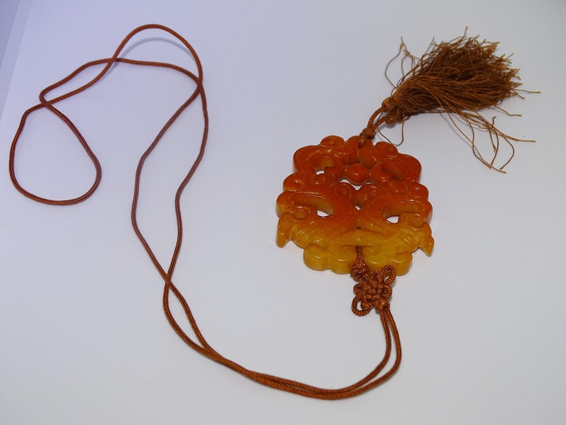 Op de afbeelding: Een gele en oranje jade hanger met ingewikkelde gravures hangt aan een bruin koord met een kwast. De hanger is met een knoop aan het koord gebonden.
