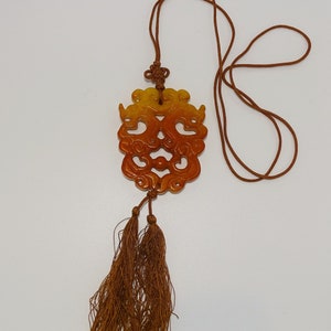 Op de afbeelding: Een bruin en geel gegraveerd jade-hanger met een kwast eraan. De hanger heeft de vorm van een draak en is bevestigd aan een bruin koord.