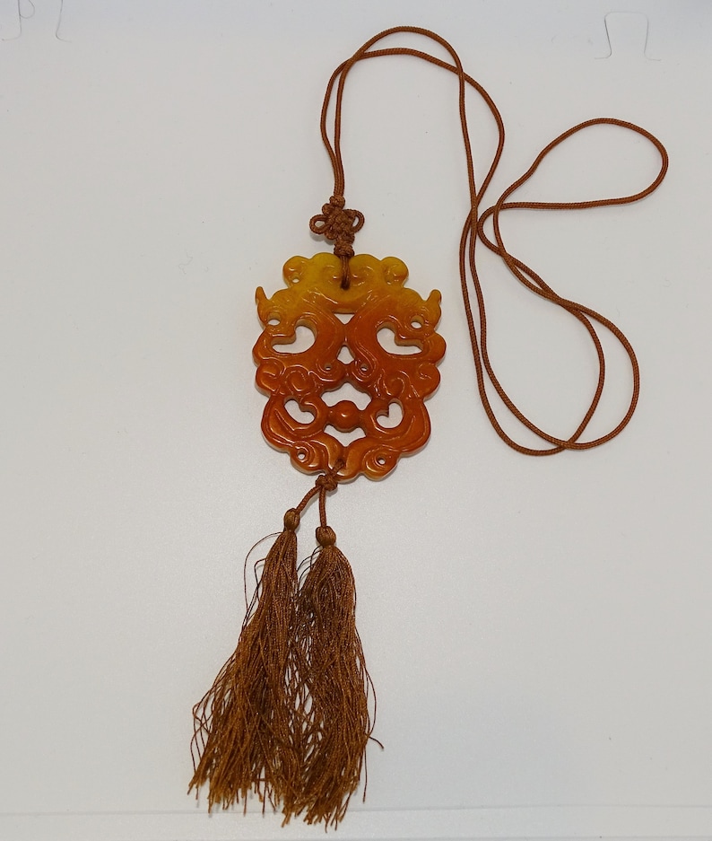 Op de afbeelding: Een bruin en geel gegraveerd jade-hanger met een kwast eraan. De hanger heeft de vorm van een draak en is bevestigd aan een bruin koord.