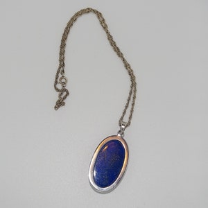Op de afbeelding: Een zilveren ketting met een blauwe ovale hanger met een lapis lazuli-steen.