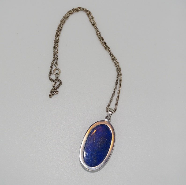 Op de afbeelding: Een zilveren ketting met een blauwe ovale hanger met een lapis lazuli-steen.