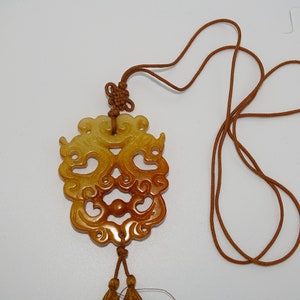 Op de afbeelding: Een ketting met een gegraveerde jade hanger, een bruin koord en kwastjes. De hanger is een gedetailleerd ontwerp van twee draken in een geel en bruin kleurenschema.
