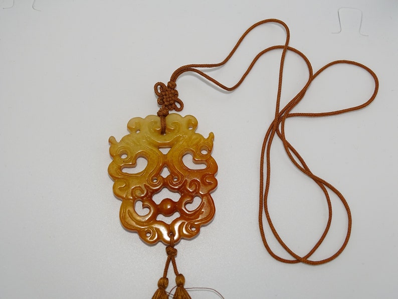 Op de afbeelding: Een ketting met een gegraveerde jade hanger, een bruin koord en kwastjes. De hanger is een gedetailleerd ontwerp van twee draken in een geel en bruin kleurenschema.